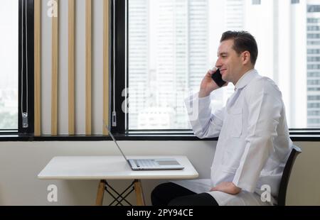 Junger weißer Arzt in weißem Kleid benutzt Handy, während er E-Mails auf einem Laptop liest. Sie saß am Fenster in der Cafeteria. Vor dem Fenster hat man einen Blick auf die Stadt mit Hochhäusern. Stockfoto