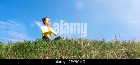 Eine Frau, die draußen auf grünem Gras sitzt und gegen den blauen Himmel meditiert. Stockfoto
