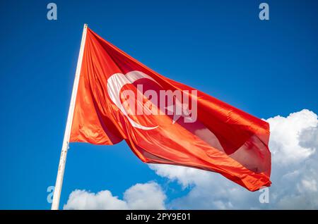 Die majestätische türkische Flagge, hoch und stolz auf einem weißen Fahnenmast, vor dem riesigen blauen Himmel mit weißen Wolken in den Ausläufern der Stockfoto