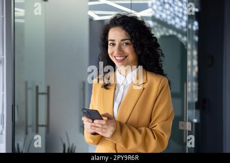 Erfolgreiche Geschäftsfrau, die im Büro arbeitet, Porträt einer Chefin in gelber Jacke und Brille, die neben dem Fenster steht, in die Kamera schaut und einen kleinen Geschäftsinhaber lächelt, ein Telefon in der Hand hält, eine Nachricht tippt Stockfoto