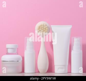 Weiße Kunststoffröhrchen und Gläser mit Creme, eine Massagebürste für die Gesichtsreinigung auf pinkfarbenem Hintergrund, Gegenstände für kosmetische Eingriffe Stockfoto