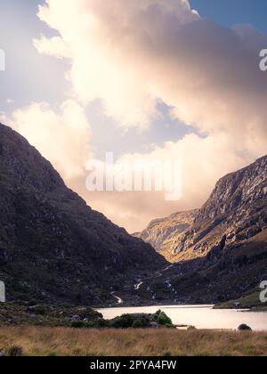 Blick auf die Gap of Dunloe in County Kerry, Irland. Die Aussicht ist das Tal hinunter mit einem See und einer Straße sichtbar. Die Sonne scheint auf den Berg Stockfoto