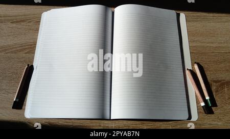 U-Turn-Notizbuch mit leeren weißen Seiten. Links und rechts neben dem Notizbuch auf dem Holztisch befinden sich farbige Bleistifte. Stockfoto