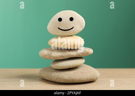 Steinstapel mit gezeichnetem fröhlichem Gesicht auf dem Tisch vor türkisfarbenem Hintergrund, Nahaufnahme. Zen-Konzept Stockfoto