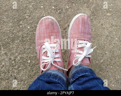 Pinkfarbene Sneaker mit weißen Schnürsenkeln an den Beinen. Blaue Jeans, darunter eingewickelt. Blick von oben. Sorgenfrei und sportlich. Stockfoto