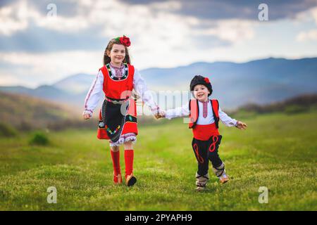 Freiheit bulgarisches Mädchen und Junge mit traditionellen ethnischen Folkloresachen, die auf einem grünen Rasen auf dem Land in Bulgarien laufen Stockfoto