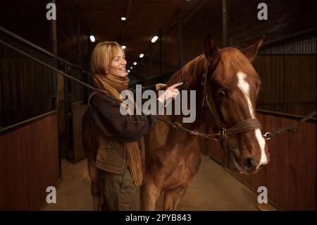 Pferderin kämmt Mähne ihres braunen Vollblüterpferdes im Stall Stockfoto