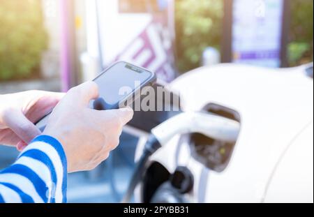 Frau, die den Smartphone-Zahlungsdienst in der mobilen App verwendet, während ein Fahrzeug mit Blur EV an der Ladestation für Elektrofahrzeuge aufgeladen wird. Ladestation für Elektrofahrzeuge. Ladestation für kommerzielle Elektrofahrzeuge. Nachhaltiger Lebensstil. Stockfoto