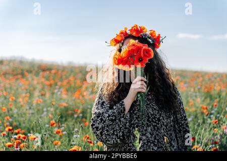Eine langhaarige, geschwungene Frau in einer Größe in einem Kranz aus rotem Mohn steht zwischen einem Feld mit Wiesenblumen und bedeckt ihr Gesicht mit einem Strauß Mohn Stockfoto