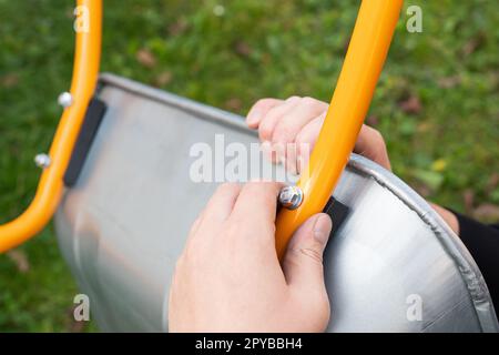 Auf der Straße steht eine Schubkarre aus Metall, ein Mann dreht eine neue Schubkarre für die Arbeit, verdreht Bolzen und Muttern. Stockfoto