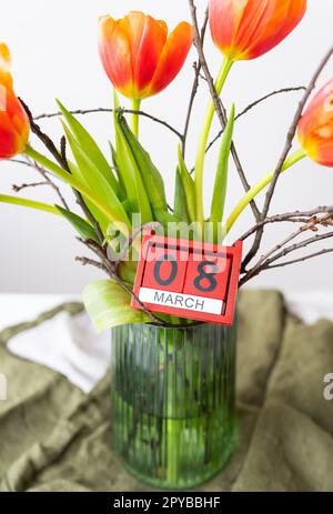 Ein sehr schöner Frühlingsstrauß in einer grünen Vase steht auf einem Tisch auf einer Tischdecke aus Leinen, orangefarbene Pfingstrosen. Holzwürfel mit der Inschrift März 8. Stockfoto