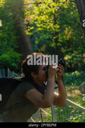 Junge afroamerikanische Fotojournalistin mit dunklen lockigen Haaren, die Fotos in einem grünen Park bei Sonnenuntergang mit der untergehenden Sonne hinter ihrem Adju macht Stockfoto