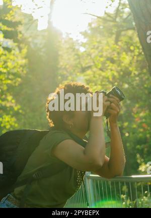 Junge afroamerikanische Fotojournalistin mit dunklen lockigen Haaren, die Fotos in einem grünen Park bei Sonnenuntergang mit der untergehenden Sonne hinter ihrem Adju macht Stockfoto