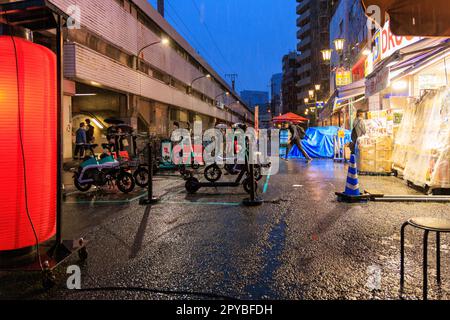 Osaka, Japan - 29. April 2023: Elektrische Roller und Fahrräder werden in regnerischer Nacht in einer hellen Apotheke und am Bahnhof geparkt Stockfoto