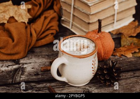 Tasse heißer weißer Kaffee auf dunklem hölzernem Hintergrund, Herbstdekorationen und Stapel Buch im Hintergrund Stockfoto