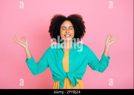 Glückliche positive schöne, trendige, lockige, afroamerikanische oder brasilianische Frau, entspannend, meditierend mit geschlossenen Augen, auf ihre geistige Gesundheit achten, lächeln, auf isoliertem rosa Hintergrund stehen Stockfoto