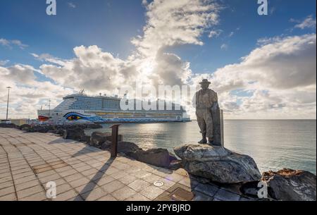 PUERTO DEL ROSARIO, SPANIEN - FEBRUAR 16: (ANMERKUNG: Für dieses Bild wurde ein abgestufter Farbfilter verwendet. Das Bild ist ein digitales Composite mit hohem Dynamikbereich.) Am 16. Februar 2023 in Puerto del Rosario, Spanien, ist ein Aida-Ozeanliner in der Nähe der Skulptur von Juan Miguel Cubas Sanchez von Pescador de Viejas im Hafen verankert. Stockfoto