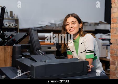 Fröhliche Frau, die in der Nähe des Druckers lächelt, während sie in der Druckerei arbeitet, Stock Image Stockfoto