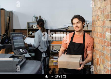 Fröhliche Arbeiterin in einer Schürze, die Kartonschachtel in der Nähe einer Kollegin in in der Druckerei hält, Stock image Stockfoto