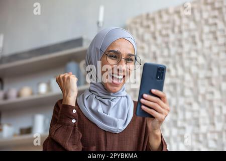 Nahaufnahme. Muslimische junge Frau im Hijab, die zu Hause steht und telefoniert. Süchtig nach Online-Spielen, platziert Wetten, hat gute Nachrichten erhalten. Freut sich und macht eine Siegesgeste mit seiner Hand. Stockfoto