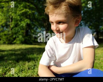 Ein süßer Junge, der lächelt und zur Seite schaut. Das Kind trägt ein weißes T-Shirt und eine blaue Hose. Blondes Haar ist ein schöner Sturz auf die Stirn. Lange blonde Wimpern. Im Hintergrund parken. Stockfoto