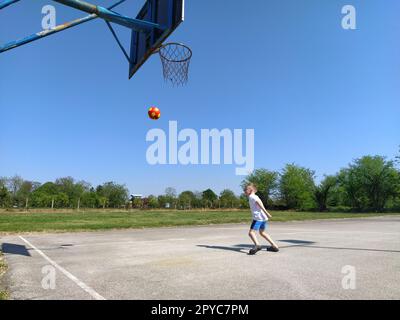 Sremska Mitrovica, Serbien. 6. Juni 2020. Ein Junge spielt Ball auf dem Spielplatz. Asphaltsportplatz. Ein Kind in einem weißen T-Shirt. Kleinkind mit blondem Haar, 7 Jahre alt. Laufen, Treten und Übungen Stockfoto