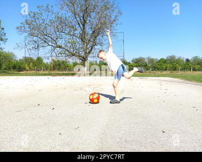 Sremska Mitrovica, Serbien. 6. Juni 2020. Ein Junge spielt Ball auf dem Spielplatz. Asphaltsportplatz. Ein Kind in einem weißen T-Shirt. Kleinkind mit blondem Haar, 7 Jahre alt. Laufen, Treten und Übungen Stockfoto