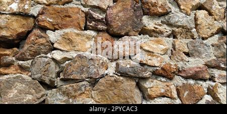 Steinmauer. Gestapelte große Ziegel und Kopfsteinpflaster in warmen Farben. Lose weiße Steine, die aus der Wandoberfläche hervorstehen. Stockfoto
