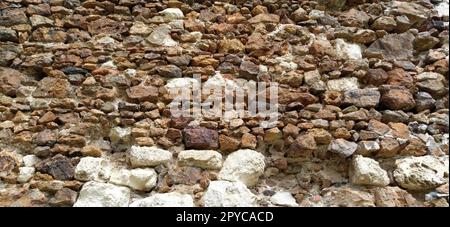 Steinmauer. Gestapelte große Ziegel und Kopfsteinpflaster in warmen Farben. Lose weiße Steine, die aus der Wandoberfläche hervorstehen. Stockfoto