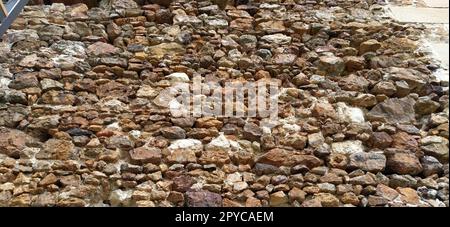 Steinmauer. Gestapelte große Ziegel und Kopfsteinpflaster in warmen Farben. Lose weiße Steine, die aus der Wandoberfläche hervorstehen. Stockfoto