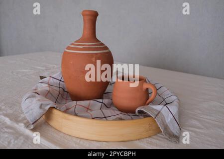 Alte, unlackierte, antike Keramikkrug aus Lehm in einem Holztablett auf einer traditionellen Tischdecke. Tonkrug und Becher zum Trinken von Wein oder Wasser. Stockfoto