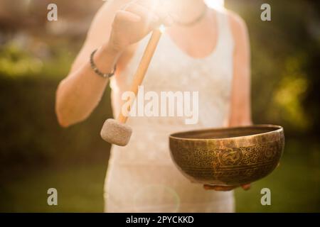 Frau spielt tibetische Bowl mit Hammer über Sunset Sky. Entspannende Musiktherapie und Sound Healing. Friedliches Meditations- und Entspannungskonzept. Stockfoto