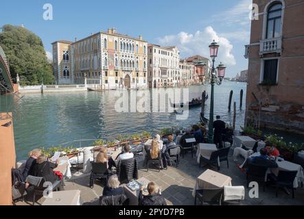 Touristen sitzen an den Tischen eines Cafés am Canale Grande in der Nähe der Rialtobrücke. Blick auf den Canal Grande von der Rialtobrücke. Venedig, Italien Stockfoto