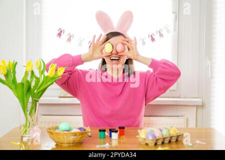 Glückliche, wunderschöne junge Frau, die lächelt und Kaninchenohren trägt und Ostereier für ihre Augen hält. Stockfoto