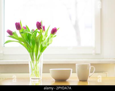 Gemütliches Frühstück mit einer Tasse heißem Tee oder Kaffee und einer Schüssel Müsli mit einem magentafarbenen Tulpenstrauß auf dem Holztisch vor einem Fenster. Guten Morgen. Speicherplatz kopieren. Hochwertiges Foto. Stockfoto