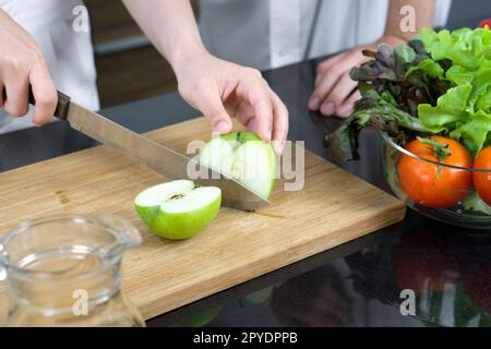 Die Hand hält ein Messer und schneidet grünen Apfel auf einem Holzbrett. Eine Glasschüssel mit verschiedenen Gemüsesorten wird auf den Küchentisch gestellt. Stockfoto