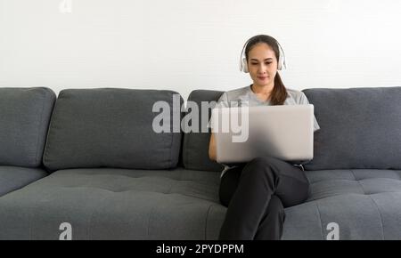 Eine asiatische Frau mittleren Alters im grauen T-Shirt hört Musik über Kopfhörer, während sie im Wohnzimmer auf einem Laptop tippt. Genießen Sie das Wochenende zu Hause. Stressfreies Konzept Stockfoto