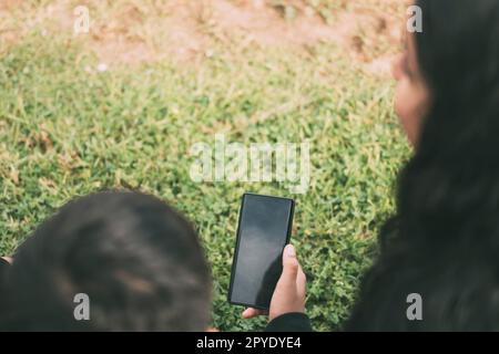 Blick von der hinteren Gruppe lachender Latinos, die auf dem Boden in einem Park mit einem Smartphone sitzen Stockfoto