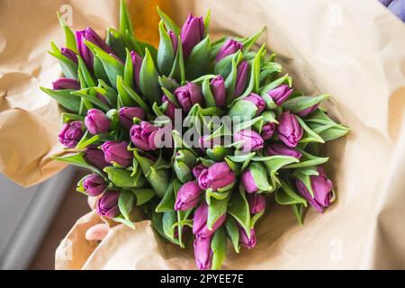 Hände wickeln lila Tulpen Bouquet in Handwerk Papier überhölzern Tisch Draufsicht. Rustikales Valentinstag-Geschenk Stockfoto