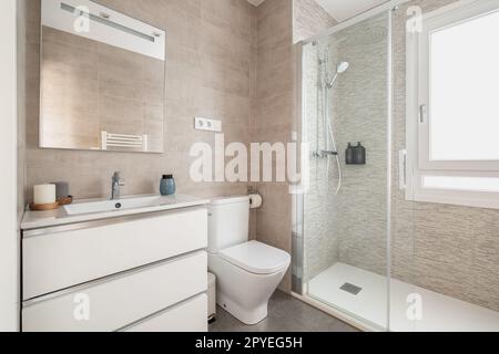 Beigefliestes Badezimmer mit gläsernen Duschtüren, Toilette und Waschbecken über Schrank mit Spiegel und Tageslicht. Modernes Badezimmerdesign Stockfoto