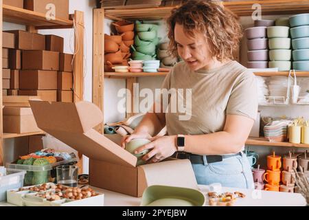 Lächeln konzentrierte, lockige Frau, die eine Sendung mit einer Babyschale aus Silikon für die Lieferung verpackt und verpackt. Arbeiten Sie zu Hause Stockfoto