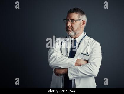 Es braucht Zeit, um an die Spitze zu kommen. Studioaufnahme eines gutaussehenden, reifen Arztes, der aufmerksam aussieht, während er mit gefalteten Armen vor einem dunklen Hintergrund steht. Stockfoto