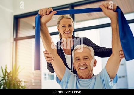 Ich führe ihn auf dem Weg zur Genesung. Eine Physiotherapeutin, die einer älteren Patientin mit einem Stretchband in ihrer Praxis hilft. Stockfoto