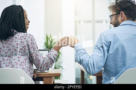 Wenn Kollegen Freunde werden. Zwei junge Kollegen, die sich in einem modernen Büro gegenseitig eine Faust an ihren Schreibtischen schlagen. Stockfoto