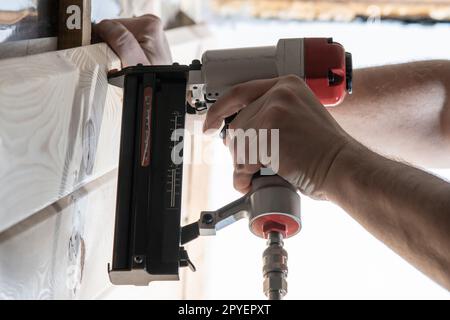 Verkürzte männliche Handschuhe verwenden eine pneumatische Tacker-Pistole zum Schneiden von Holzbrettern. Kratzer an Metallnägeln und Heftklammern Stockfoto