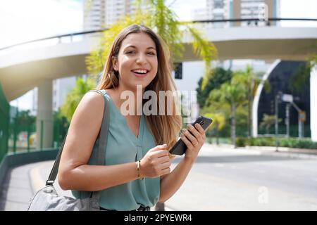 Halblanges Porträt einer fröhlichen brasilianischen Frau auf der Straße mit Smartphone. Ein wunderschönes Millennials-Mädchen, das in der Stadt in der Hand eines Mobiltelefons in die Kamera schaut. Stockfoto