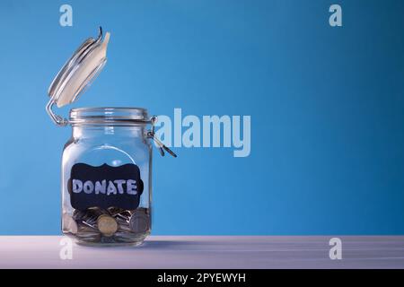 Glasbehälter mit Münzen und Kreideetikett Spenden auf blauem Hintergrund. Spende- und Wohltätigkeitskonzept. Speicherplatz kopieren. Stockfoto