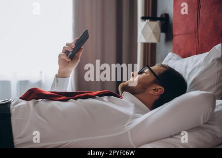 Ein Geschäftsmann ruht auf einem Bett in einem Hotelzimmer. Ein junger Mann benutzt sein Smartphone, während er sich ausruht. Stockfoto