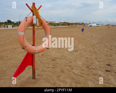 Anapa, Russland, 10. August 2021 Lifebuoy mit einer roten Flagge an einem Sandstrand. Orangefarbenes Rettungsboot an einer Stange, um Menschen zu retten, die im Meer ertrinken. Rettungspunkt am Ufer. Stürmisches Wetter am Schwarzen Meer Stockfoto