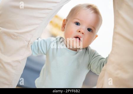 Porträt eines niedlichen neugierigen kleinen Jungen, das zu Hause die ersten Schritte unternimmt und an der Hose seines Vaters festhält. Süßer kleiner Junge, der Laufen lernt. Stockfoto